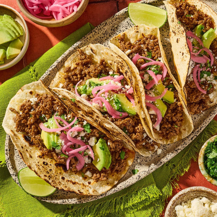 A platter of three street tacos filled with ground beef seasoned with Cholula Medium Taco Seasoning Mix 1 Oz and topped with avocado slices and pickled red onions.