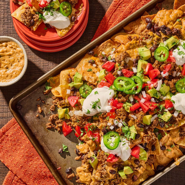 A sheet pan of loaded beef nachos is topped with fresh avocado, tomatoes, jalapeños, and sour cream, all seasoned with Cholula Medium Taco Seasoning Mix 1 Oz.