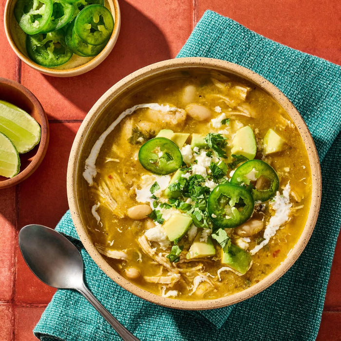 A hearty bowl of white chicken chili prepared with Cholula Medium Taco Seasoning Mix 1 Oz is garnished with fresh avocado, sliced jalapeños, crumbled cheese, and a swirl of sour cream.