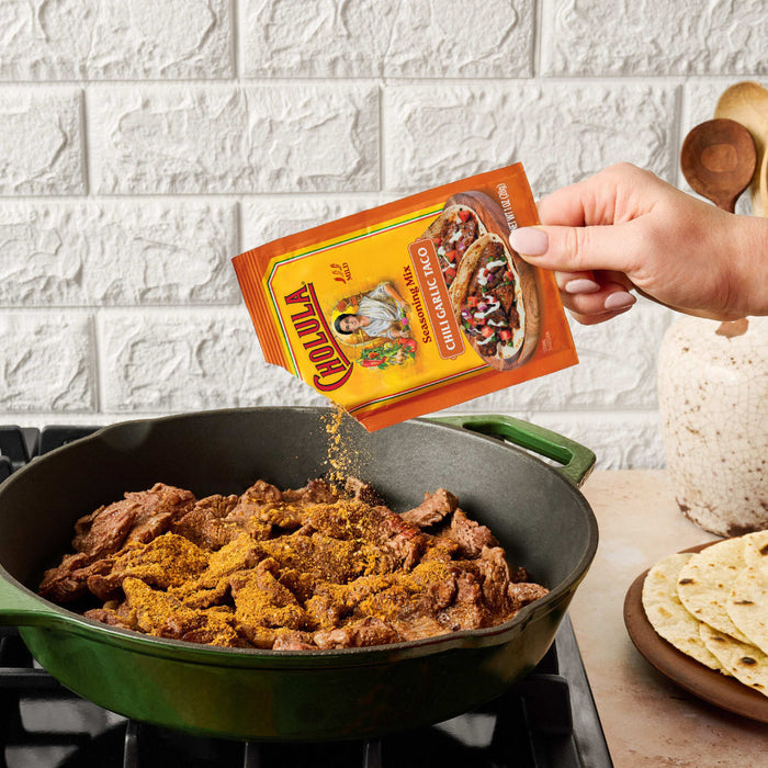 A hand pours Cholula Medium Chili Garlic Taco Seasoning Mix 1 Oz over pieces of steak browning in a green cast iron skillet on a stovetop.