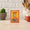 A packet of Cholula Medium Chili Garlic Taco Seasoning Mix 1 Oz stands on a kitchen counter surrounded by fresh limes, garlic, and wooden cooking utensils.