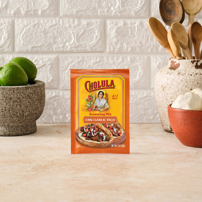 A packet of Cholula Medium Chili Garlic Taco Seasoning Mix 1 Oz stands on a kitchen counter surrounded by fresh limes, garlic, and wooden cooking utensils.