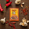 A packet of Cholula Medium Chili Garlic Taco Seasoning Mix 1 Oz is displayed on a rustic wooden surface surrounded by fresh red chili peppers, whole garlic bulbs, and dried chili flakes.