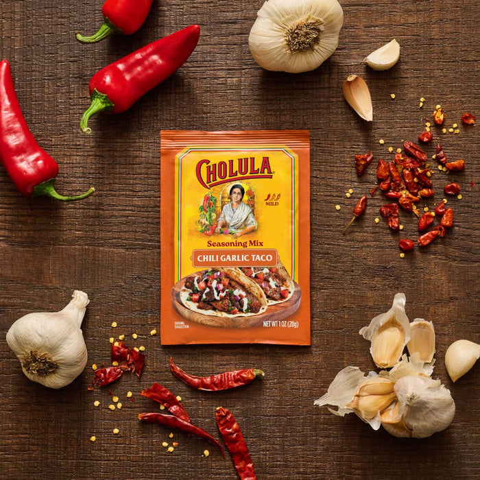 A packet of Cholula Medium Chili Garlic Taco Seasoning Mix 1 Oz is displayed on a rustic wooden surface surrounded by fresh red chili peppers, whole garlic bulbs, and dried chili flakes.