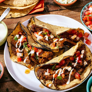 Four flavorful tacos, potentially seasoned with Cholula Chili Gar Taco Seas Mix 1 Oz, are presented on a white plate, garnished with fresh pico de gallo and a creamy drizzle, alongside extra tortillas and side dishes.
