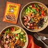 Two steak burrito bowls topped with avocado and salsa are served alongside a packet of Cholula Seasoning Mix Chili Garlic Taco.