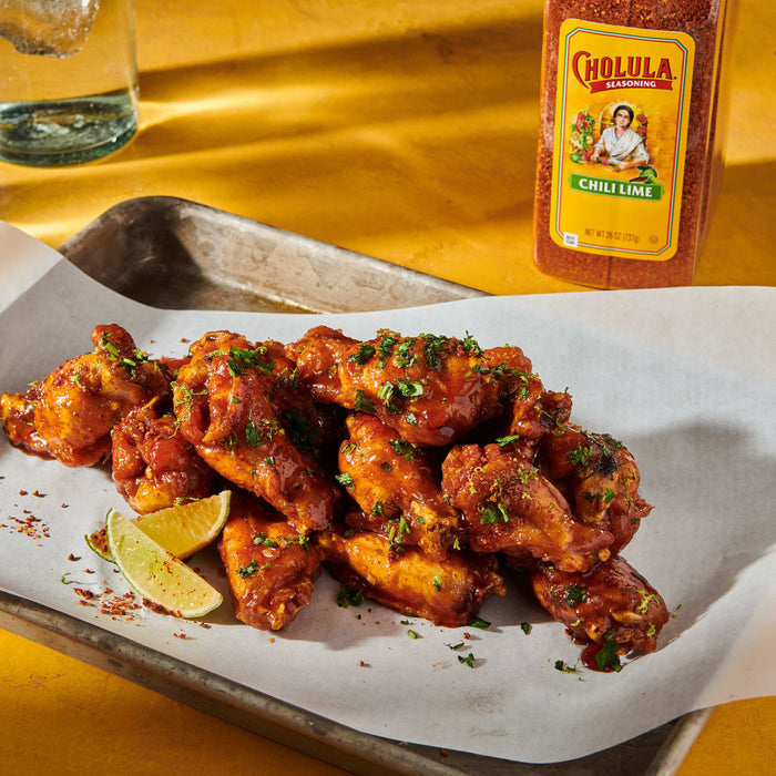 A plate of chicken wings with lime wedges and a bottle of Cholula Chili Lime seasoning.