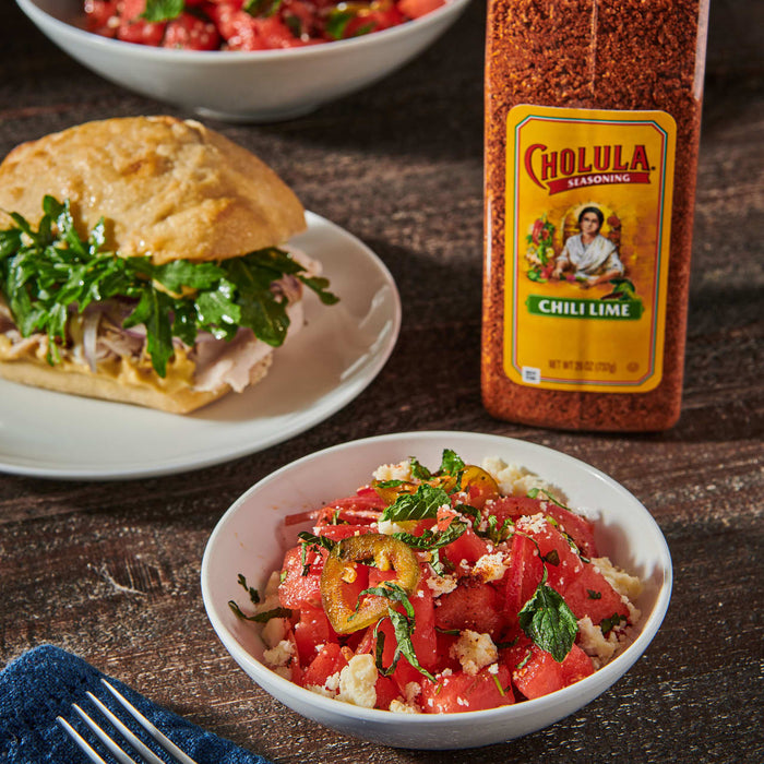 A bowl of watermelon salad with feta cheese, jalapeno peppers, and mint, next to a sandwich and a bottle of Cholula Chili Lime seasoning.