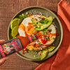 A hand holds a bottle of Cholula Hot Sauce over a plate of eggs, avocado, and tortilla chips.