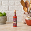 A bottle of Cholula Extra Hot Hot Sauce sits on a countertop with limes, garlic, and wooden spoons.