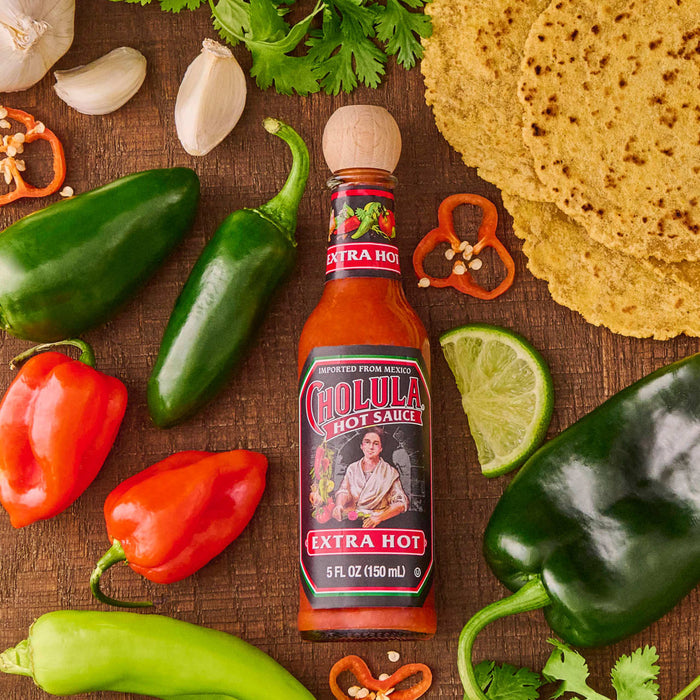 A bottle of Cholula Extra Hot sauce with peppers, limes, and tortillas.