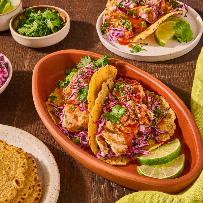 Two tacos filled with fish, cabbage, and sauce are on a plate with lime wedges.