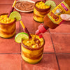 A hand pours Cholula Chamoy Sauce into a mango drink with a chili-rimmed glass and a lime wedge.