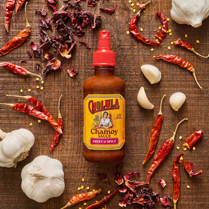 A bottle of Cholula Chamoy Sweet &amp; Spicy sauce is surrounded by dried chili peppers, garlic cloves, and hibiscus flowers on a wooden surface.