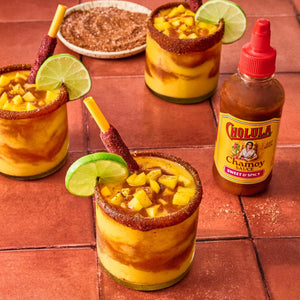 Three glasses of mango chamoyada with lime wedges and straws, a dish of chili powder, and a bottle of Cholula Chamoy Sauce are arranged on a red tile surface.