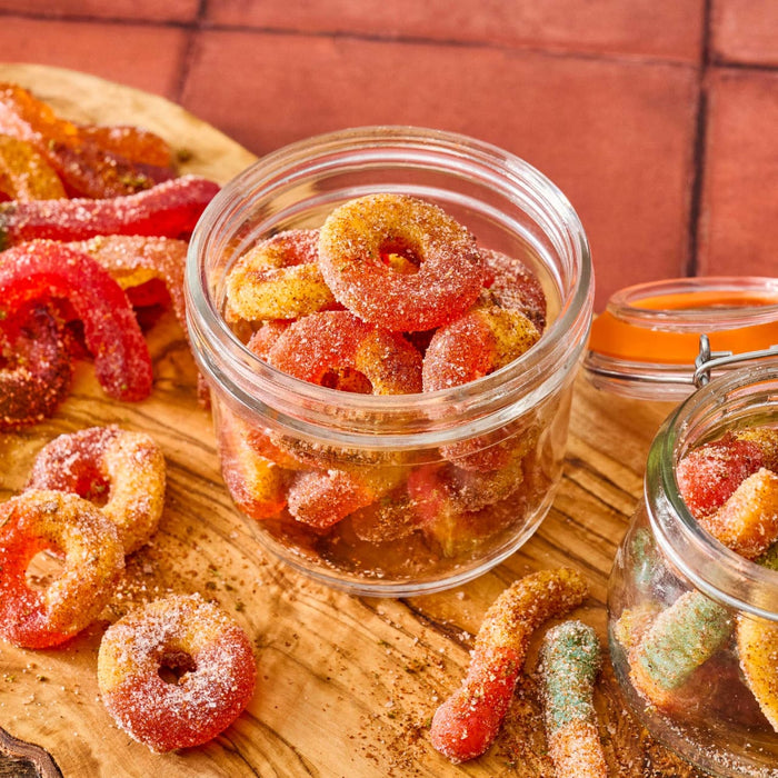 A close-up shot of gummy peach rings and gummy worms covered in sugar and chili powder, displayed in glass jars and on a wooden board.