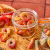 A close-up shot of gummy peach rings and gummy worms covered in sugar and chili powder, displayed in glass jars and on a wooden board.