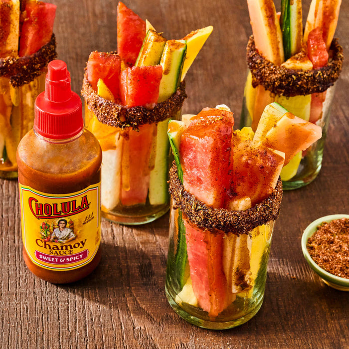 This is an image of Cholula Chamoy Sweet &amp; Spicy sauce next to glasses filled with fruit and vegetables, with the rims of the glasses coated in a dark spice.