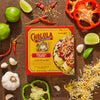 A Cholula Enchilada Sauce box is surrounded by fresh ingredients like limes, peppers, garlic, and shredded cheese, suggesting a recipe or meal preparation.