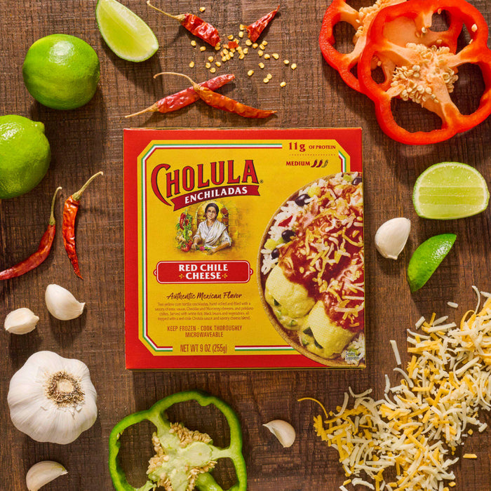 A Cholula Enchilada Sauce box is surrounded by fresh ingredients like limes, peppers, garlic, and shredded cheese, suggesting a recipe or meal preparation.