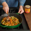 A person is adding Cholula salsa to a green cast iron skillet filled with vegetables.