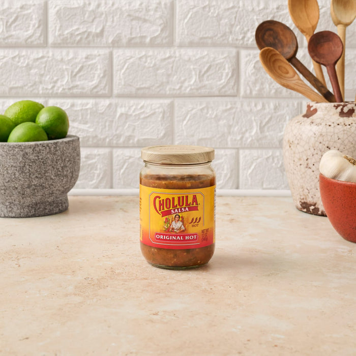 A jar of Cholula Original Hot Sauce sits on a countertop next to limes and wooden spoons.