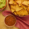 A top-down view of a bowl of guacamole, a bowl of salsa, and a bowl of tortilla chips, arranged on a red cloth.