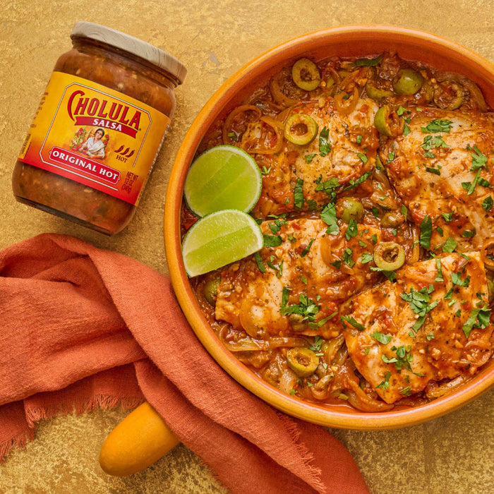 A jar of Cholula Original Hot salsa sits next to a dish of fish with salsa, olives, and lime wedges.