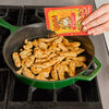 A hand pours Cholula Original Fajita seasoning mix into a green pan with cooked chicken.