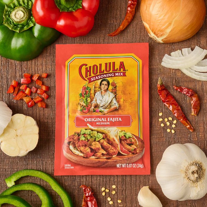 A Cholula Original Fajita Seasoning Mix packet is surrounded by fresh ingredients like peppers, onions, and garlic.