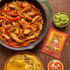 A skillet of chicken fajitas with Cholula seasoning mix, guacamole, pico de gallo, and tortillas on a wooden table.