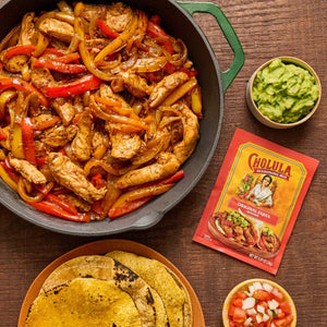 A skillet of chicken fajitas with Cholula seasoning mix, guacamole, pico de gallo, and tortillas on a wooden table.