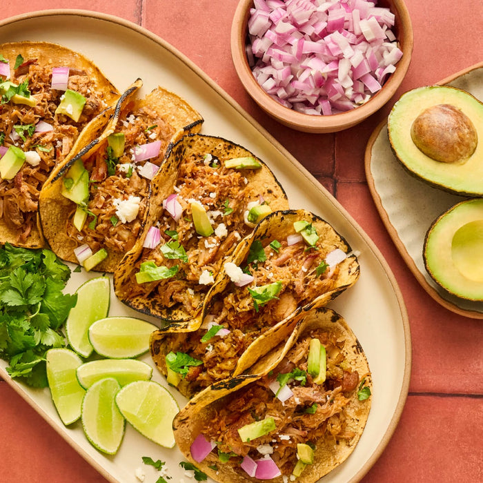 A platter of delicious tacos topped with shredded meat, avocado, red onion, and cilantro, accompanied by lime wedges and a bowl of diced red onion, showcasing a vibrant and appetizing meal.
