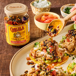 A jar of Cholula Chili Crisp 8 Oz sits on a wooden table next to a plate of tostadas, with a hand spooning the chili crisp sauce over one of the loaded tostadas.