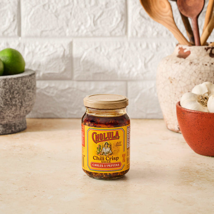 A jar of Cholula Chili Crisp Sauce sits on a counter with limes garlic and wooden spoons in the background.