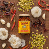 A jar of Cholula Chili Crisp 8 Oz, labeled Chiles &amp; Pepitas, sits on a wooden surface surrounded by dried red chilies, garlic bulbs, cloves, and green pumpkin seeds.