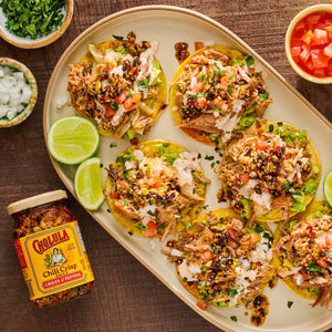 A platter of six loaded tostadas with shredded meat, guacamole, and fresh toppings, accompanied by a jar of Cholula Chili Crisp 8 Oz.