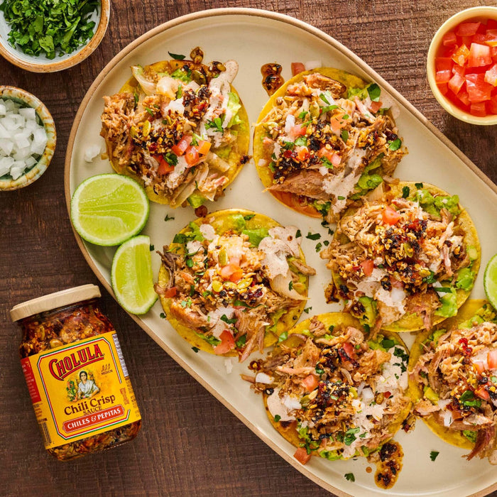 A platter of six loaded tostadas with shredded meat, guacamole, and fresh toppings, accompanied by a jar of Cholula Chili Crisp 8 Oz.
