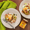 Two plates of avocado toast topped with fried eggs and Cholula Chili Crisp sauce are displayed on a wooden table with a green napkin and a jar of the sauce.