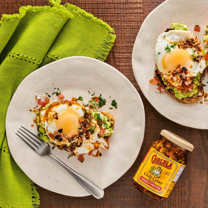 Two plates of avocado toast topped with fried eggs and Cholula Chili Crisp sauce are displayed on a wooden table with a green napkin and a jar of the sauce.