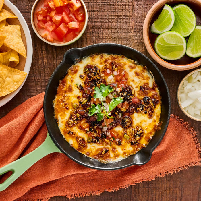 A cast iron skillet of melted cheese dip, generously topped with Cholula Chili Crisp 8 Oz, diced tomatoes, and cilantro, served alongside tortilla chips, lime wedges, and diced onions on a wooden table.