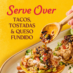 A hand pours Cholula Chili Crisp 8 Oz from a wooden spoon onto a taco, highlighting its versatility for tacos, tostadas, and queso fundido.