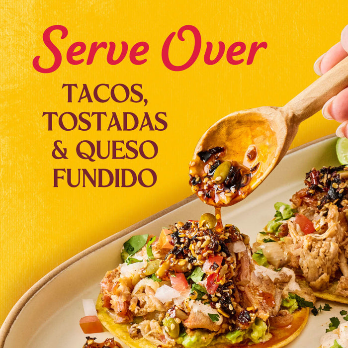 A hand pours Cholula Chili Crisp 8 Oz from a wooden spoon onto a taco, highlighting its versatility for tacos, tostadas, and queso fundido.