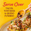 A wooden spoon pours a savory sauce over tacos tostadas and queso fundido showcasing a delicious culinary creation.