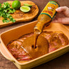 A hand pours Cholula Carne Asada Marinade &amp; Cooking Sauce over raw meat in a baking dish with limes and cilantro in the background.