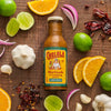 A bottle of Cholula Carne Asada Marinade &amp; Cooking Sauce is surrounded by limes oranges red onions garlic and dried chiles on a wooden surface.