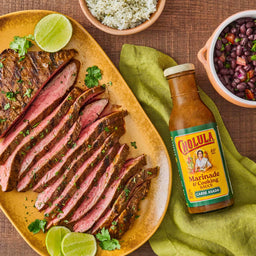 A plate of sliced carne asada with lime and cilantro accompanied by rice black beans and Cholula Marinade &amp; Cooking Sauce.