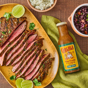 A plate of sliced carne asada with lime and cilantro accompanied by rice black beans and Cholula Marinade &amp; Cooking Sauce.