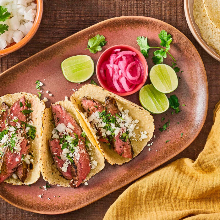 Overhead shot of a plate of steak tacos with lime wedges pickled onions and a bowl of diced onions.