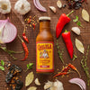 A bottle of Cholula Birria Cooking &amp; Simmer Sauce is surrounded by ingredients like garlic red onion red peppers and bay leaves on a wooden surface.